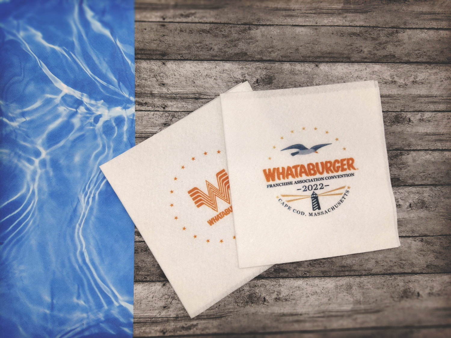 A wooden pier above clear blue water. Two napkins lay on top with one reading "Whataburger Franchise Association Convention 2022" written above a lighthouse, and under a seagull. The second napkin underneath is the Whataburger logo in a circle of stars.