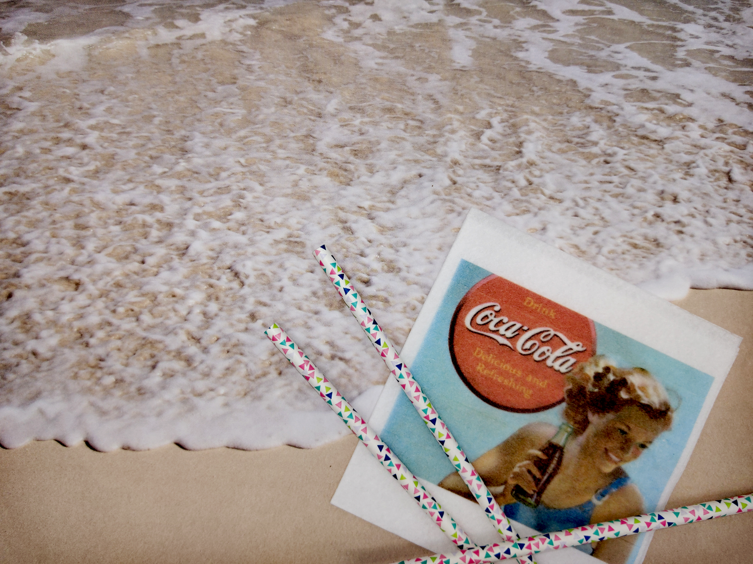 A sandy beach with waves washing up to the shore. A napkin with three colorful straws lays in the foreground, with a full-color print of a mid-1900s Coca-Cola advertisement printed on top.